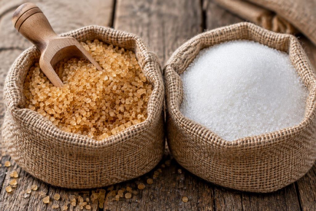 Deux sacs en toile de jute ouverts contenant du sucre roux et du sucre blanc cristallisé posés sur une table en bois avec une pelle en bois.